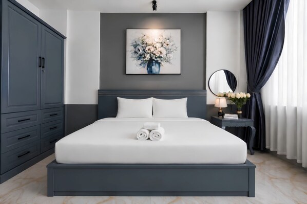 Une chambre d’hôtel moderne avec un grand lit, une armoire bleu foncé, une table de chevet avec une lampe et des fleurs, et un tableau accroché au mur.