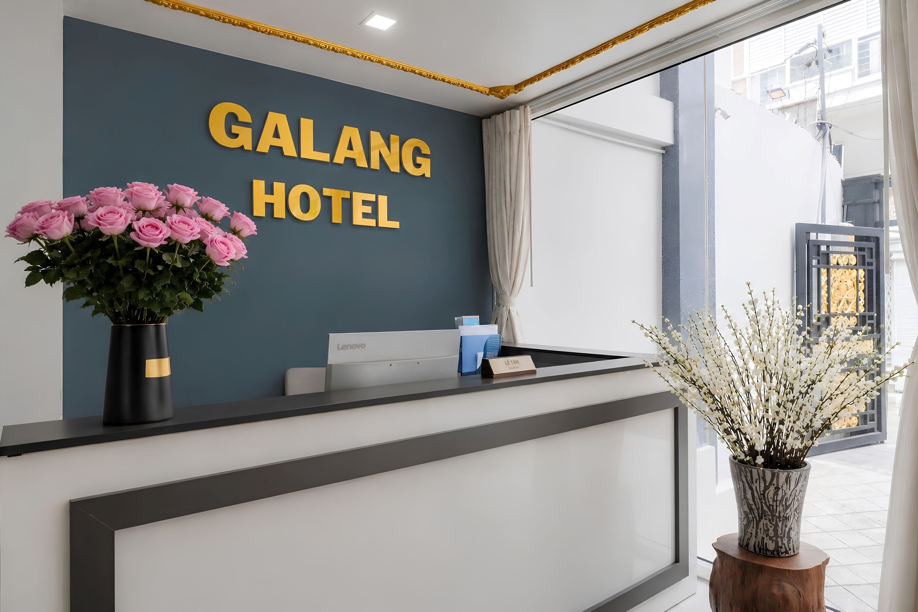 A hotel reception desk with a dark wall, a sign reading 'GALANG HOTEL', a vase of pink roses, and a desk with a nameplate.