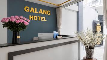 A hotel reception desk with a dark wall, a sign reading 'GALANG HOTEL', a vase of pink roses, and a desk with a nameplate.