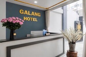 Un comptoir d’accueil d’hôtel avec un mur sombre, une enseigne indiquant « HÔTEL GALANG  », un vase de roses roses et un bureau avec une plaque nominative.