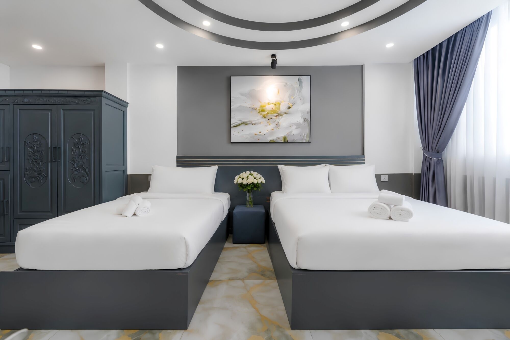 A hotel room with two beds, a large artwork above the beds, and a wardrobe with floral carvings.