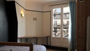 A bedroom with a bed, a desk, a window with a view of buildings, and a door with a curtain. - Lovely bright and quiet 2-room apartment, Marais. 5th floor. (Paris)