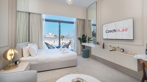 A modern hotel room with a large bed, a TV mounted on the wall, a balcony with a view of the city, and a desk with a lamp and a clock.