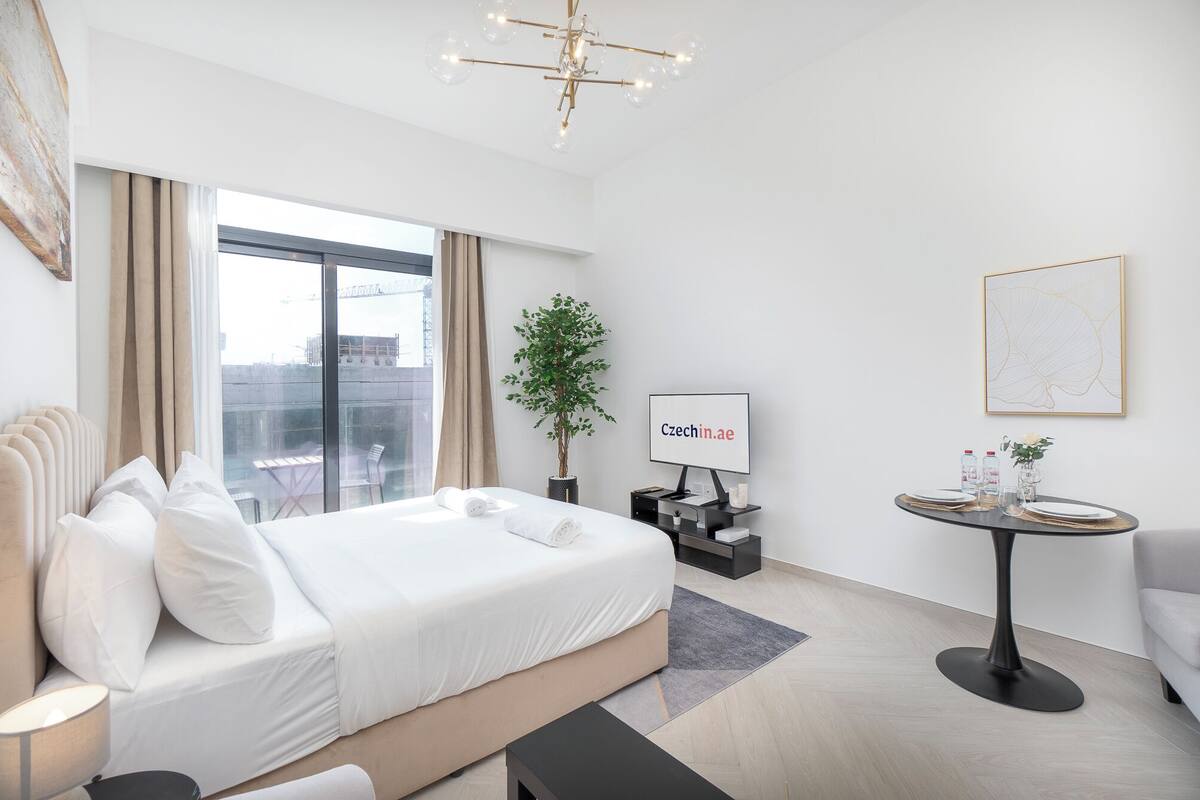 A modern hotel room with a large bed, a desk, a chair, a small table, a potted plant, and a city view.