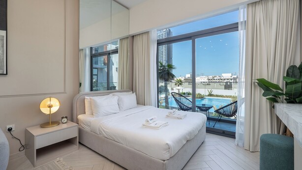A hotel room with a bed, bedside table, lamp, and a view of a swimming pool and buildings.