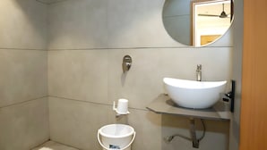 A modern bathroom with a large mirror, a white sink, and a white bucket. - Hotel Rio Orbit (Rajkot)