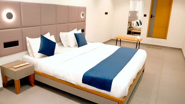 A modern hotel room with a large bed, bedside tables, a nightstand, a wooden bench, and a mirror. - Hotel Rio Orbit (Rajkot)