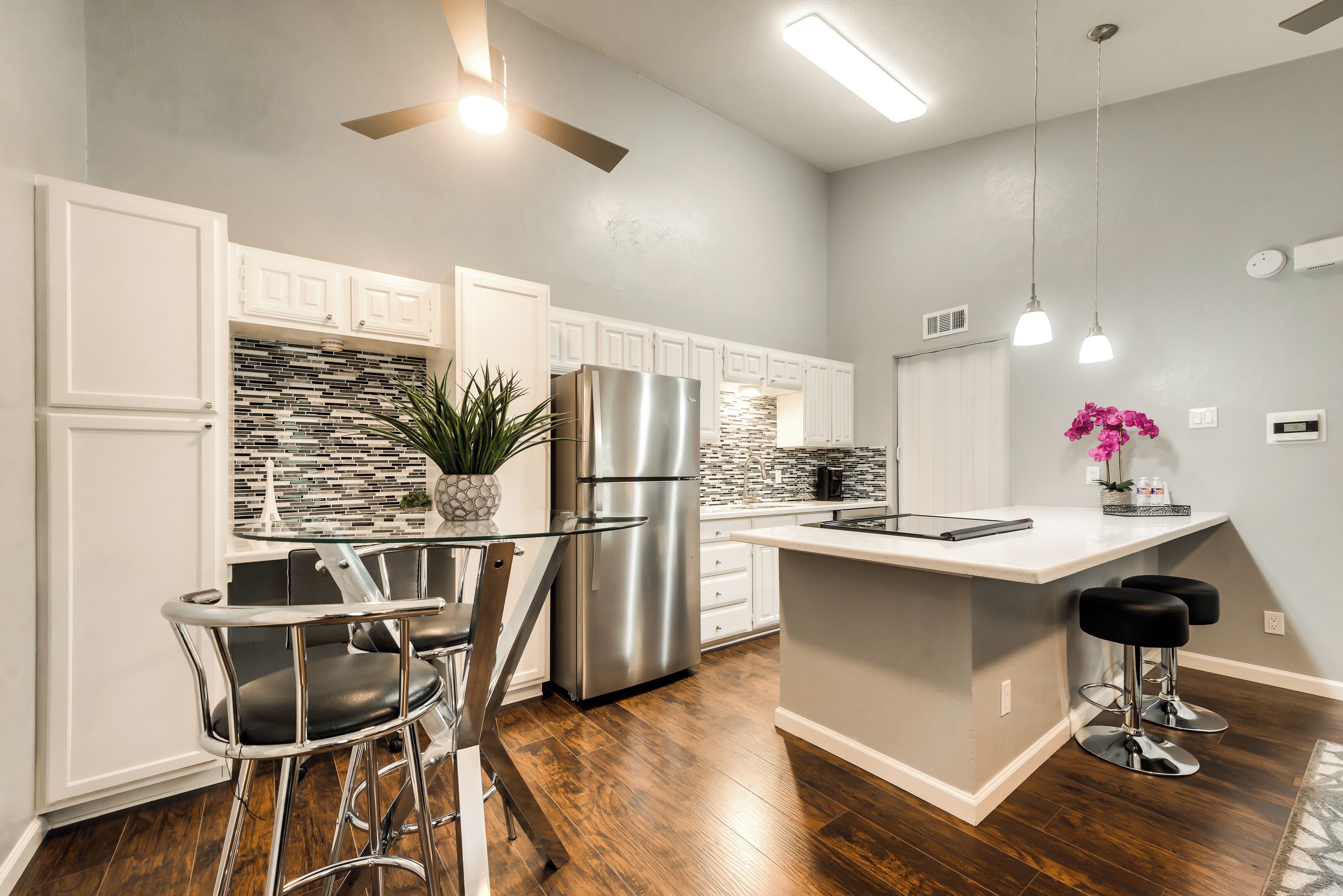 A modern kitchen with white cabinets, a stainless steel refrigerator, a glass-top island with bar stools, and a ceiling fan.