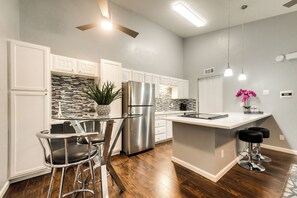 A modern kitchen with white cabinets, a stainless steel refrigerator, a glass-top island with bar stools, and a ceiling fan.