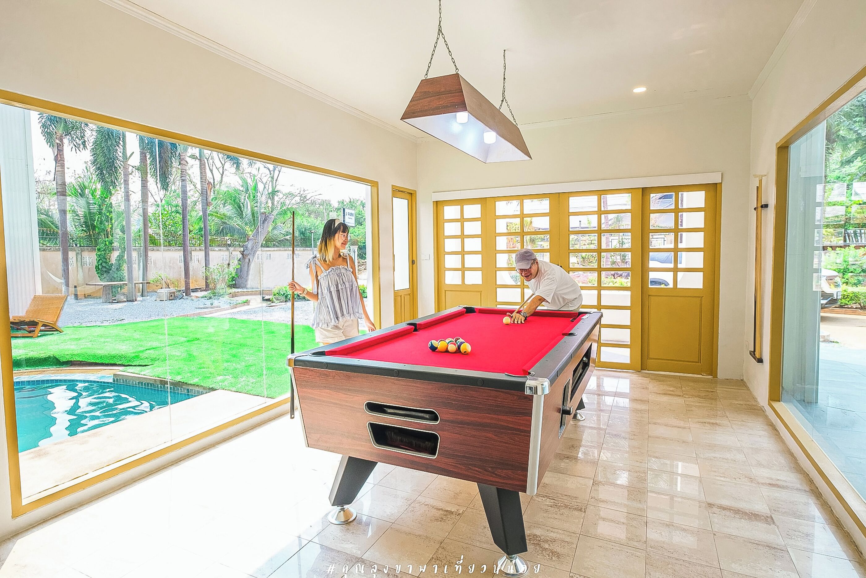 A pool table in a room with large windows and a view of a swimming pool and greenery outside.