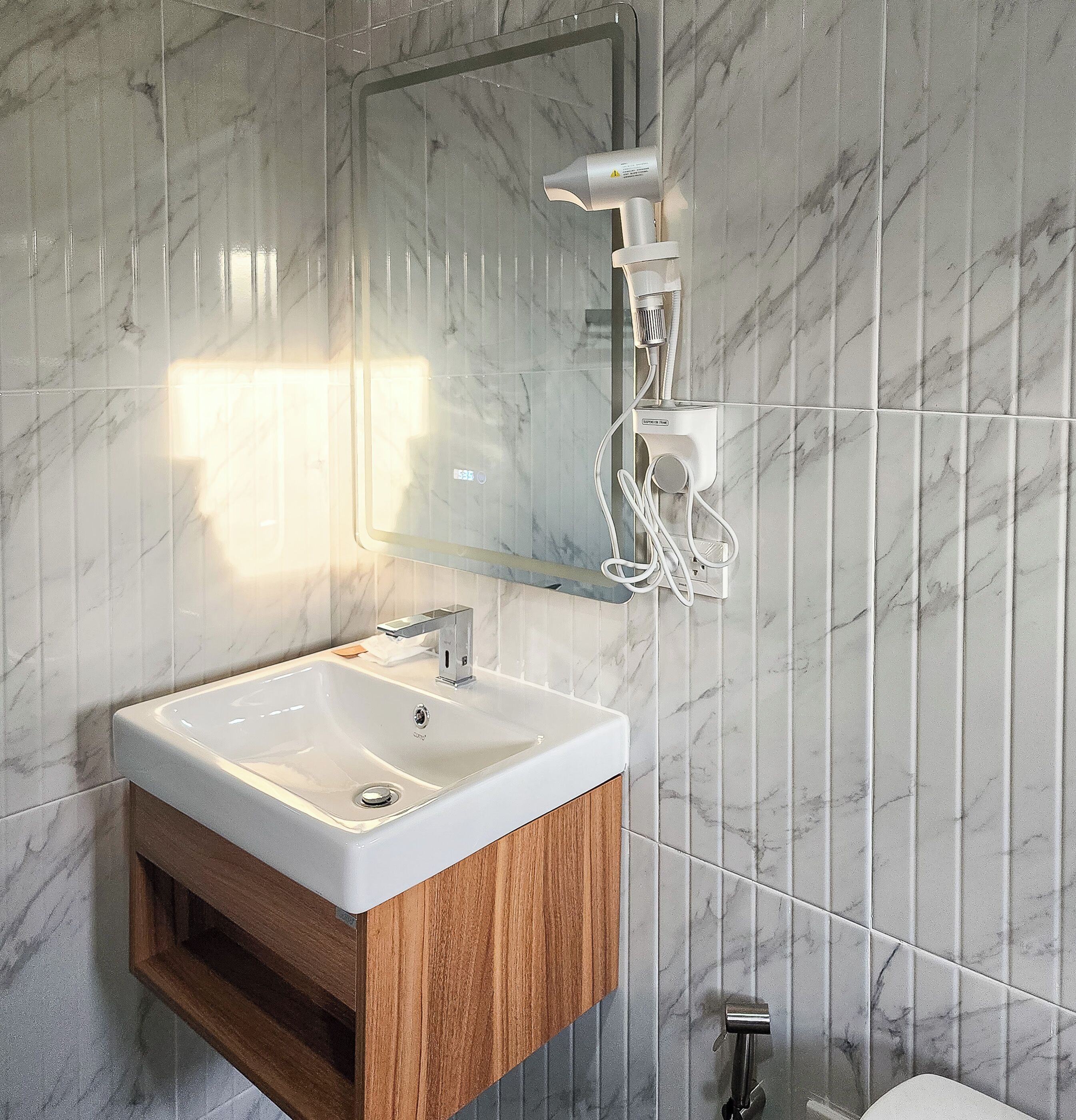 Ein Badezimmer mit Marmorwand, einem weißen Waschbecken mit Holzschrank, einem Spiegel und einer Handbrause.