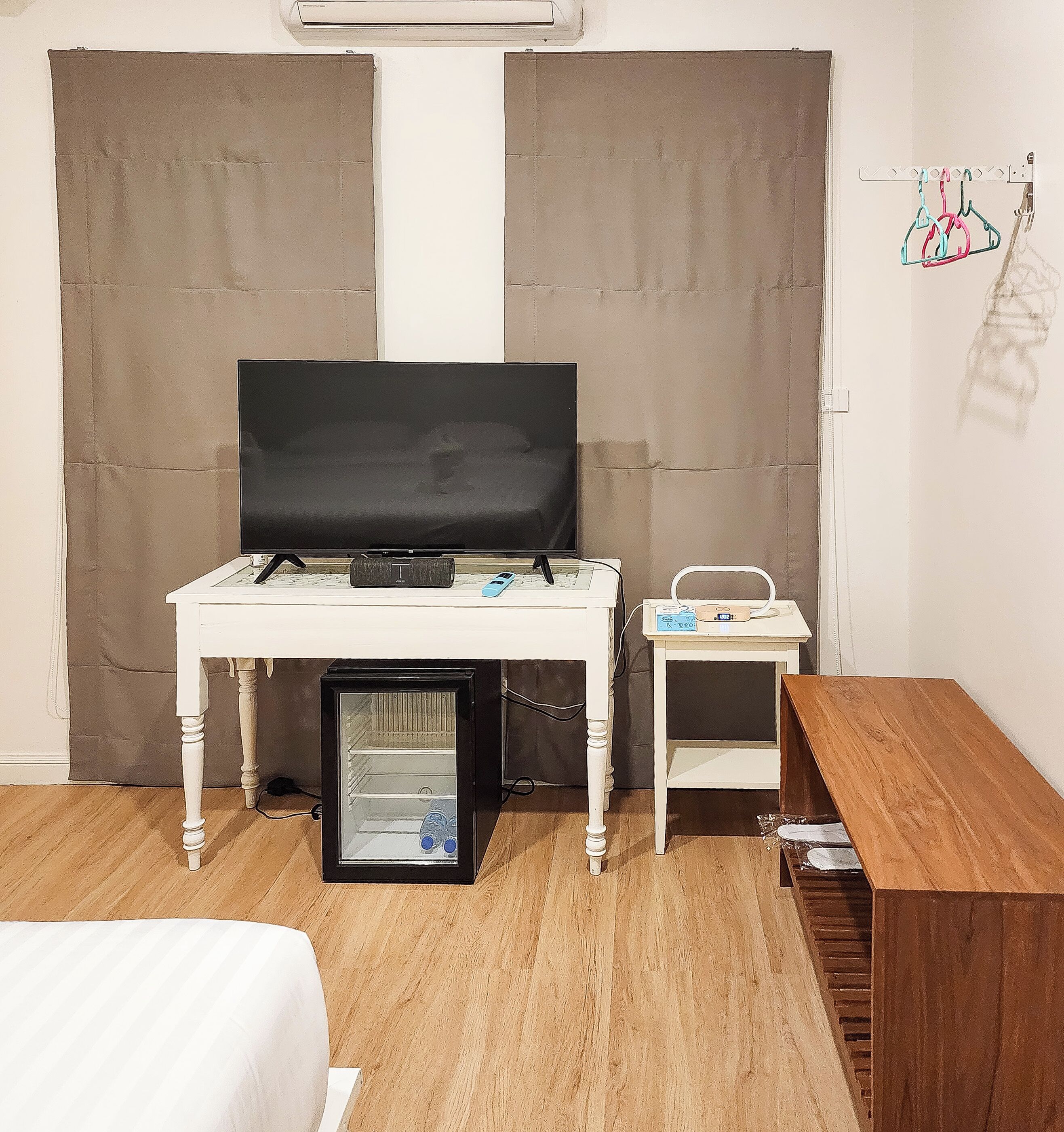 Ein kompaktes Zimmer mit Fernseher auf einem weißen Tisch, einem kleinen Beistelltisch mit Radio, einer Minikühlschrank und einer Holzbank.