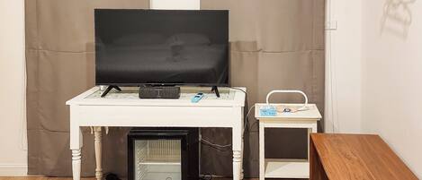 Ein kompaktes Zimmer mit Fernseher auf einem weißen Tisch, einem kleinen Beistelltisch mit Radio, einer Minikühlschrank und einer Holzbank.