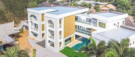 A modern multi-story building with a swimming pool, surrounded by lush greenery and palm trees.