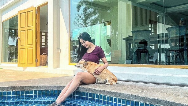 Una mujer sentada junto a una piscina con un perro, frente a un edificio moderno con grandes ventanales.
