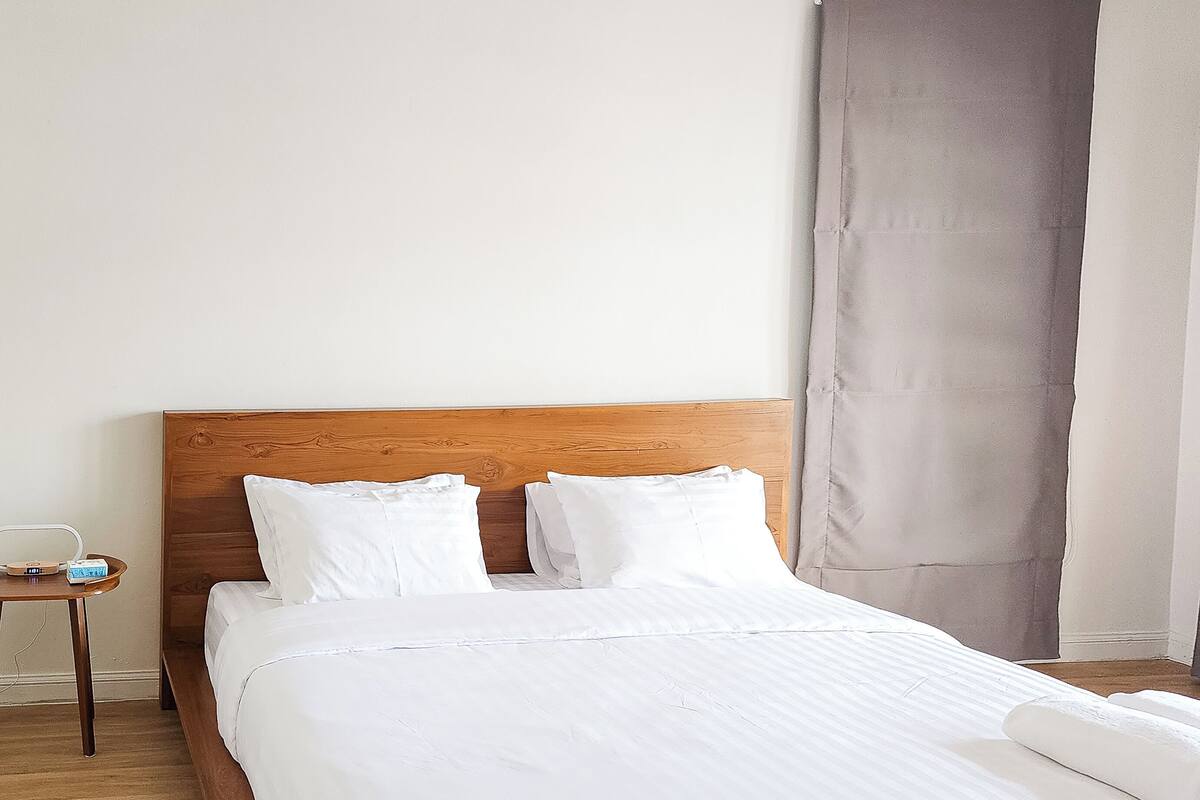 Una cama con cabecera de madera, ropa de cama blanca y una mesita de noche con lámpara.