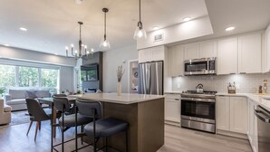 A modern kitchen with stainless steel appliances, a central island with bar stools, and a dining area with a sofa and chairs. - Serenity at Sparrowhawk Lodge Brand New 2Bd 2Ba (Dead Man's Flats)