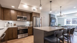Une cuisine moderne avec des meubles en bois, des appareils en acier inoxydable, un îlot central doté de tabourets de bar et un éclairage suspendu.