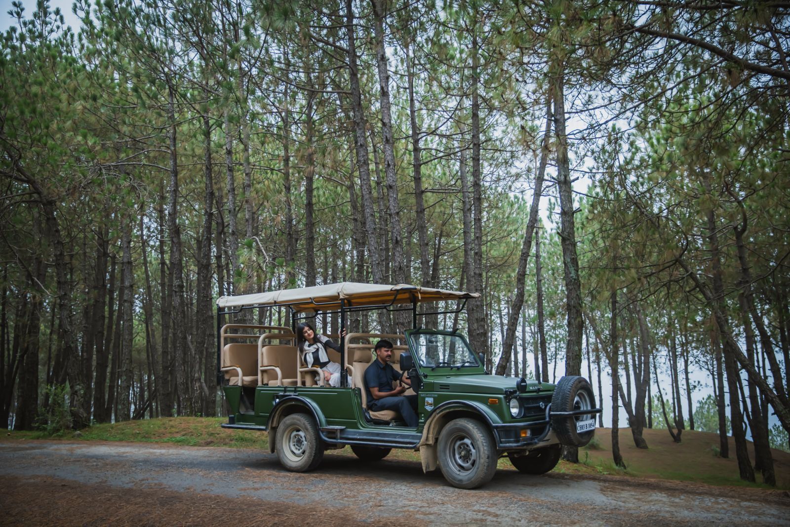 Un vehículo safari verde con toldo, dos pasajeros y un conductor, viajando por un camino forestal.