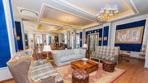 A spacious living room with a large sofa, two armchairs, a coffee table, and a decorative ottoman. The room features a chandelier, a large mirror, and a painting on the wall. - The Manor Wellness & Sport Hotel (Shimla)