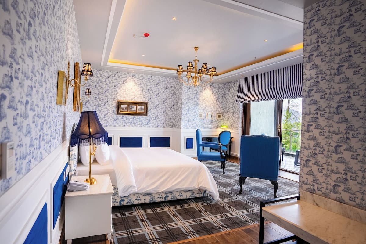 A bedroom with a large bed, a desk, and a chair. There is a chandelier, a window, and a door leading to a balcony.