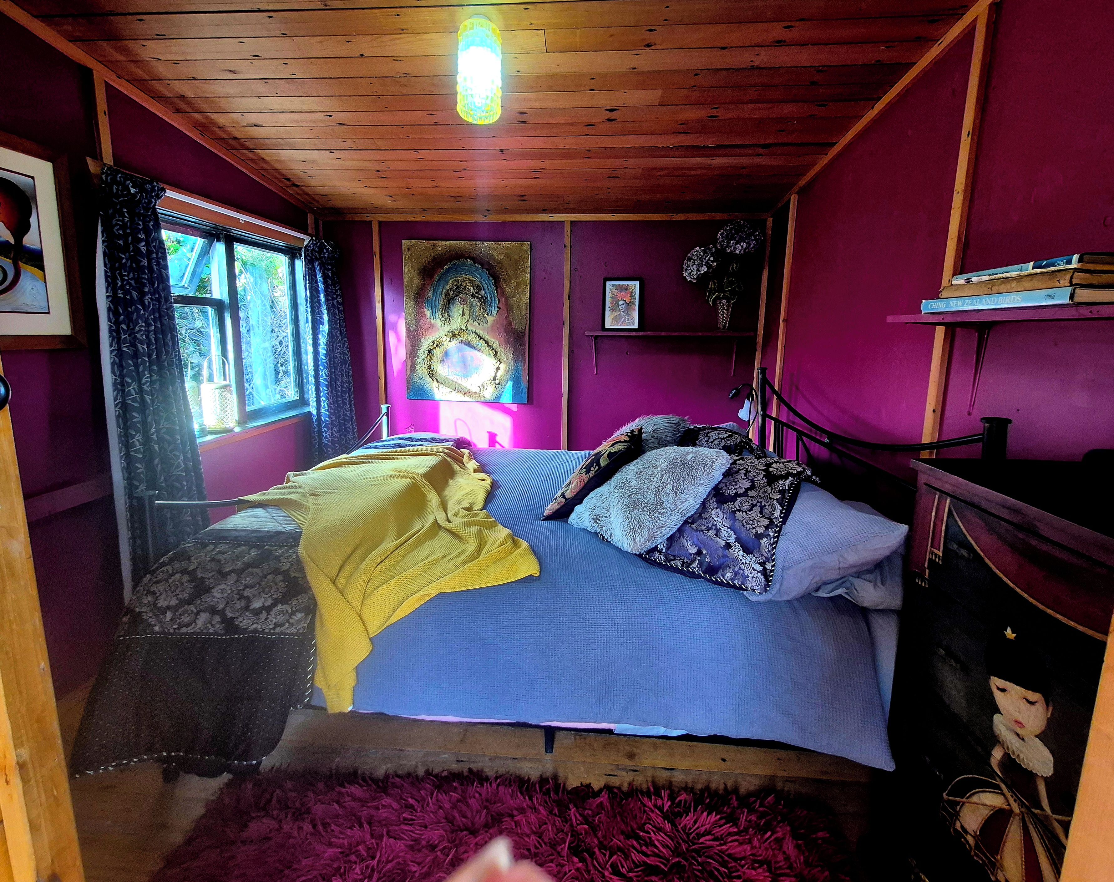 A room with a bed, a window with curtains, a painting on the wall, and a shelf with books.
