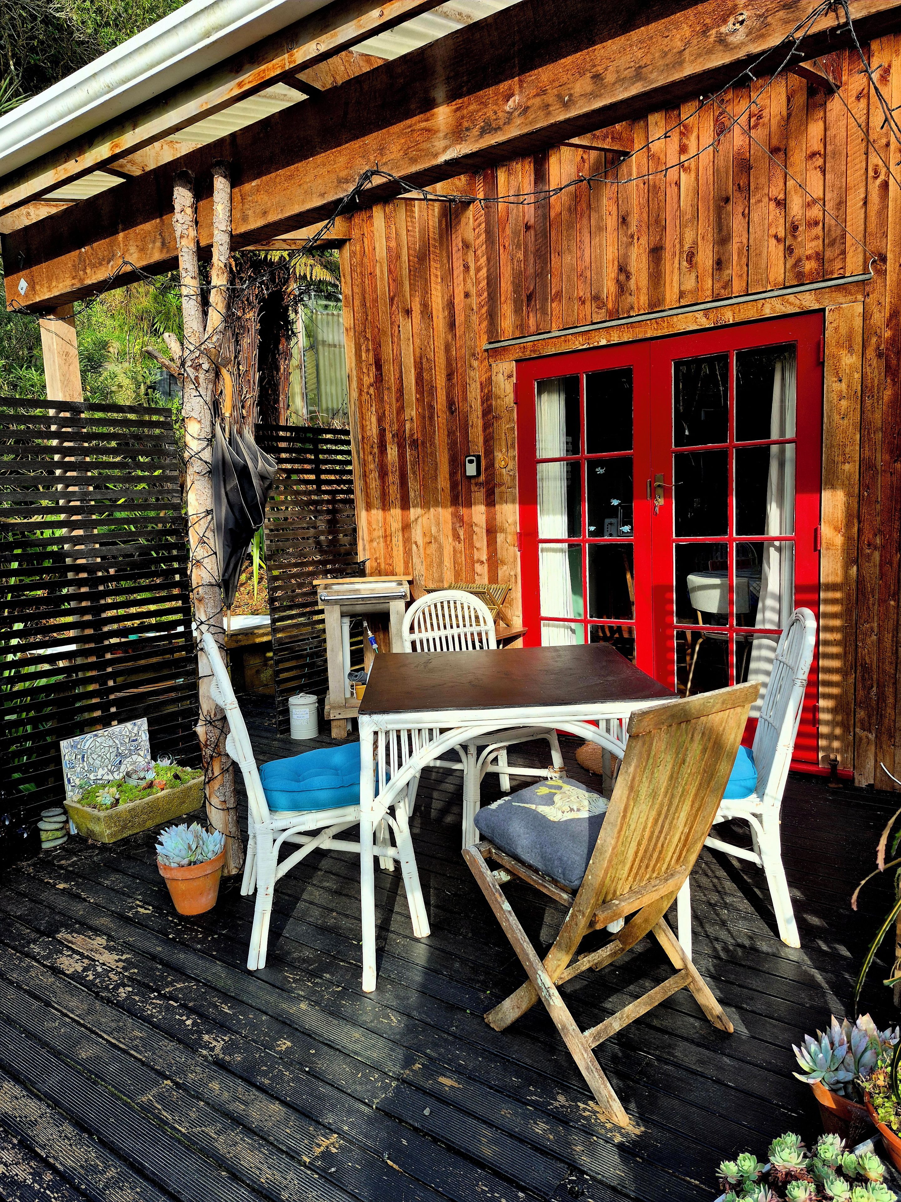 Una terraza exterior de madera con una mesa y sillas, una puerta roja y una valla de madera.