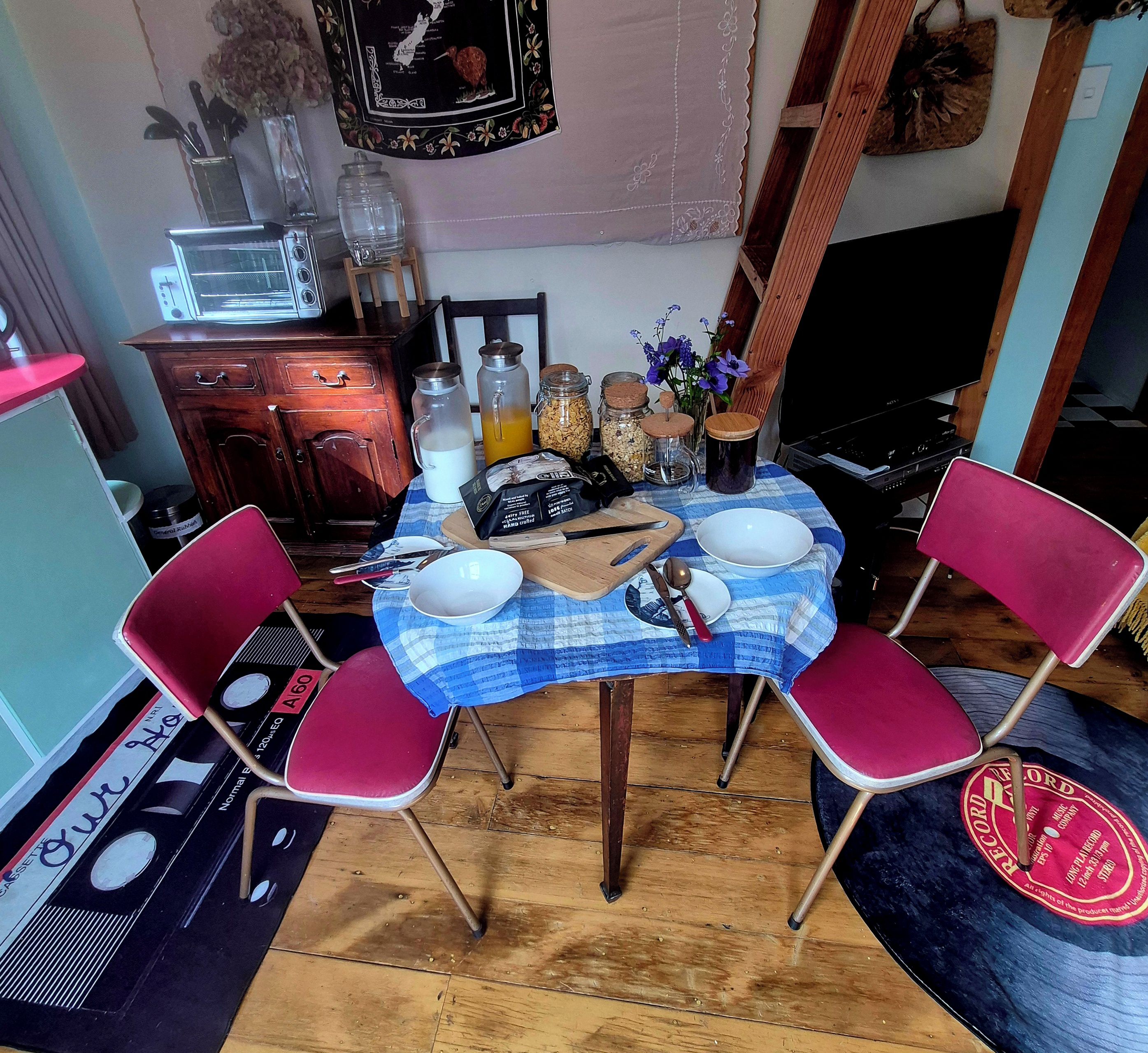 Ein Essbereich mit einem Tisch für zwei Personen, umgeben von roten Stühlen, einer blauen Tischdecke und verschiedenen Küchengegenständen.