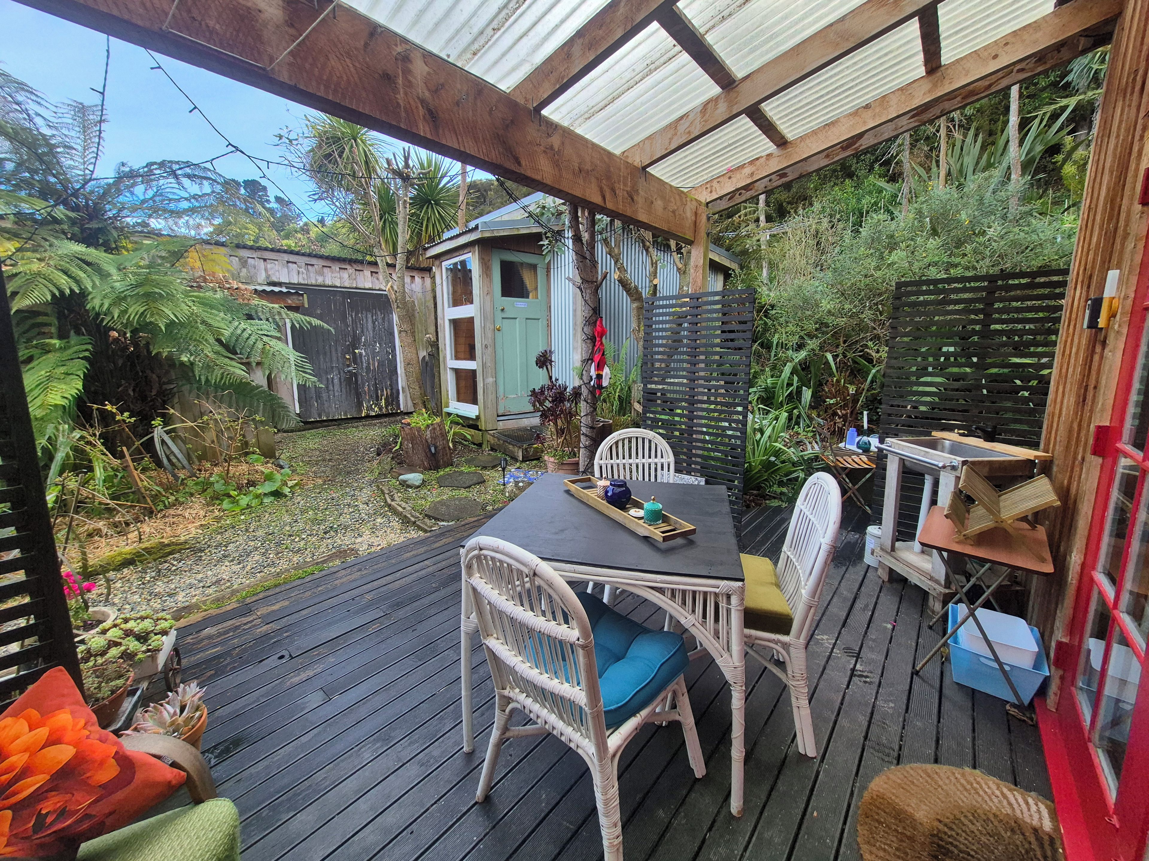 Une terrasse extérieure couverte, dotée d’une table et de chaises, entourée de verdure et agrémentée d’un petit abri de jardin.