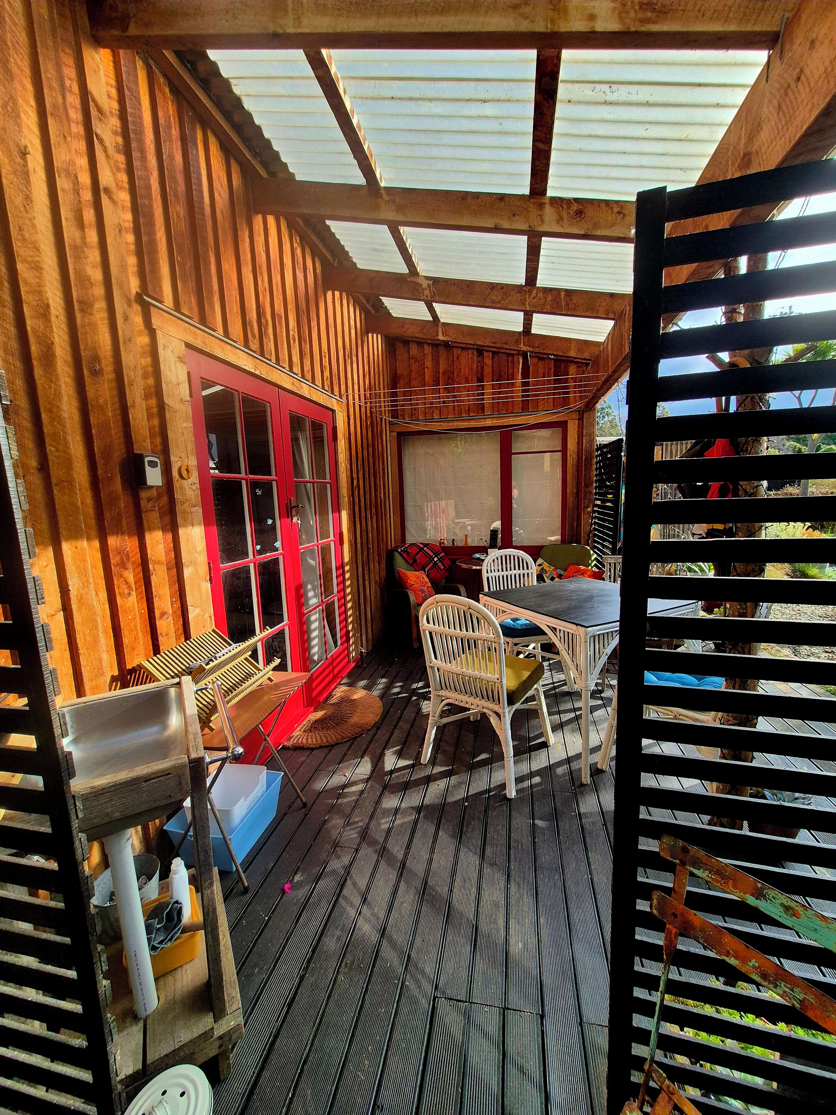 Une terrasse en bois avec une table et des chaises, une porte rouge et une fenêtre donnant sur l’extérieur.