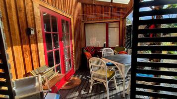 Une terrasse en bois avec une table et des chaises, une porte rouge et une fenêtre donnant sur l’extérieur.