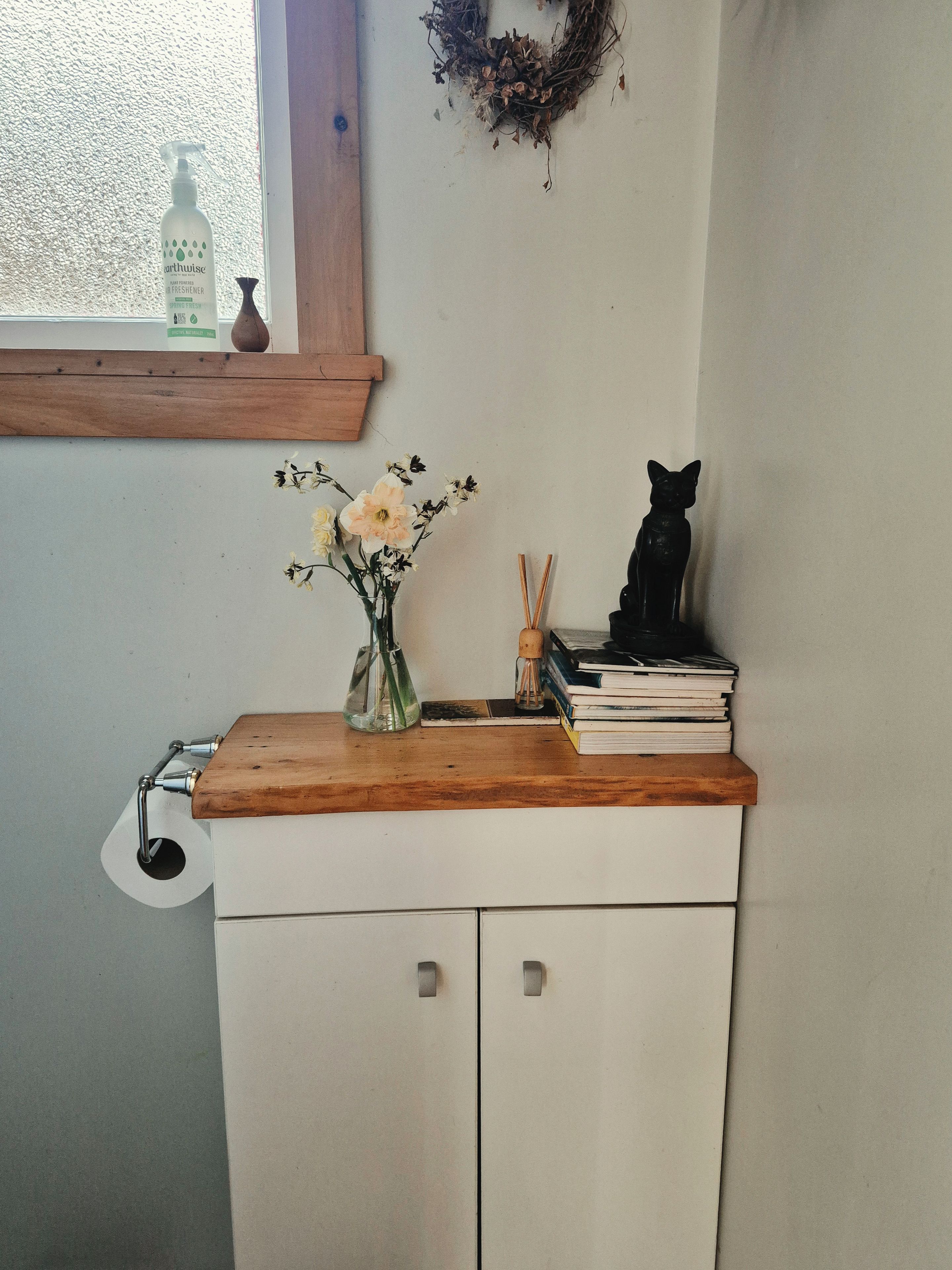 A bathroom with a wooden vanity, a vase with flowers, a black cat figurine, and a window with a wreath.