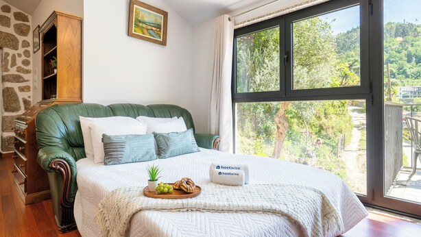 A bedroom with a green upholstered headboard, a bed with white bedding, a bedside table with a tray of fruit, and a window with a view of greenery.
