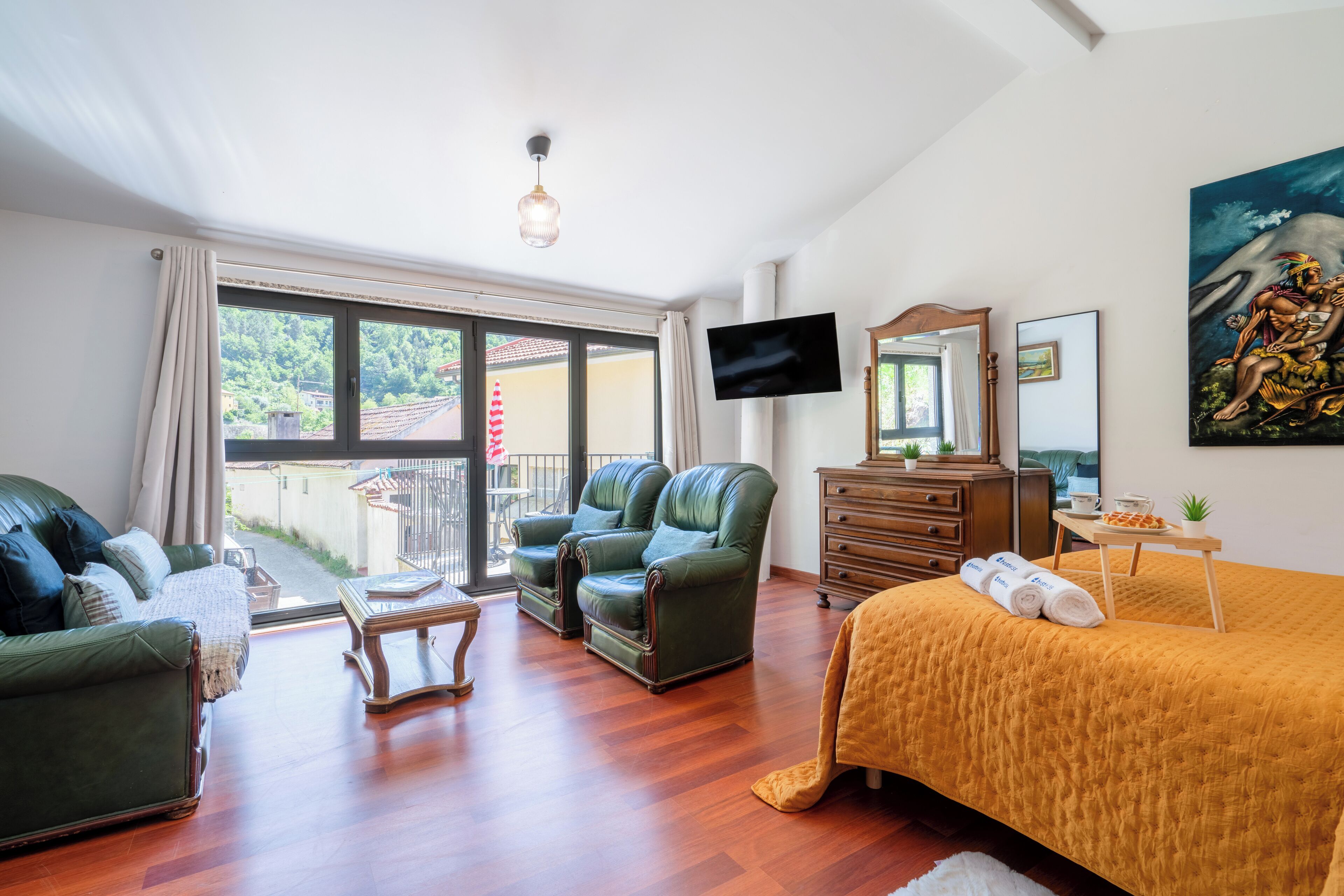 A hotel room with a bed, two green leather chairs, a wooden dresser, a television, and a balcony with a view of greenery.