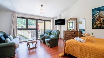 A hotel room with a bed, two green leather chairs, a wooden dresser, a television, and a balcony with a view of greenery.