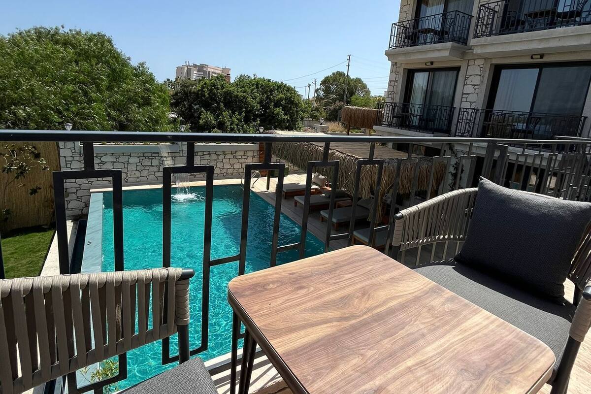 A rooftop pool area with a table and chairs, a stone wall, and a building with balconies in the background.