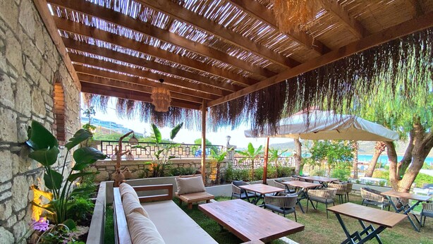 An outdoor patio area with wooden benches and tables, surrounded by greenery and a stone wall. - Lotus Port  (Cesme)