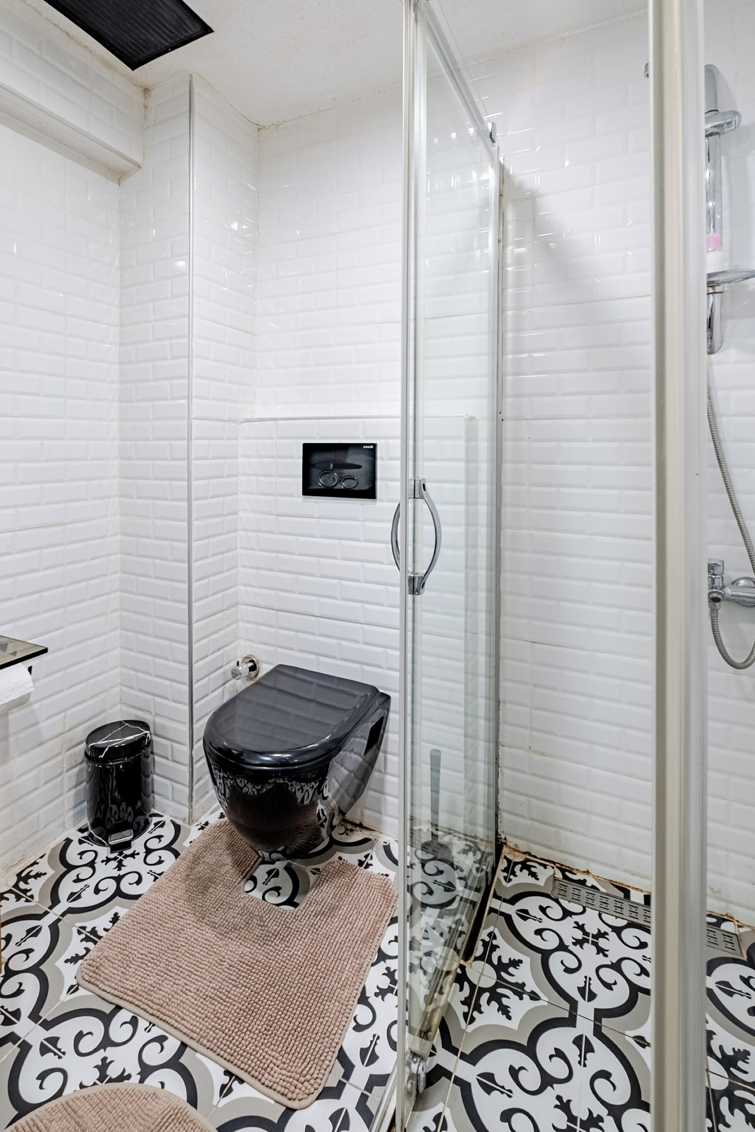 Ein Badezimmer mit weißen Fliesen an den Wänden, einer schwarzen Toilette, einer Duschkabine aus Glas und fliesenverziertem Boden.