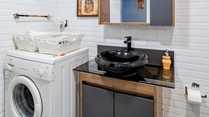 A compact bathroom with a washing machine, a sink with a black basin, a wooden cabinet, and a mirror above the sink. - Cozy Central Flat w/Netflix & Elevator Near Metro 2 minute walking distance (İstanbul)