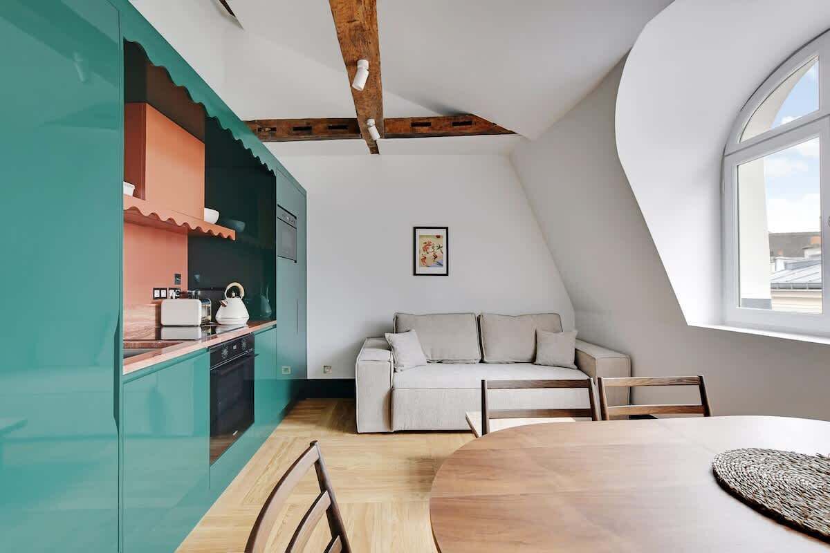 A modern kitchen with teal cabinetry, a wooden dining table, and a white sofa in a loft-style room with a sloped ceiling and large windows.