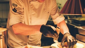 Un chef preparando comida en una cocina con superficies de acero inoxidable y una lámpara colgante de cobre.