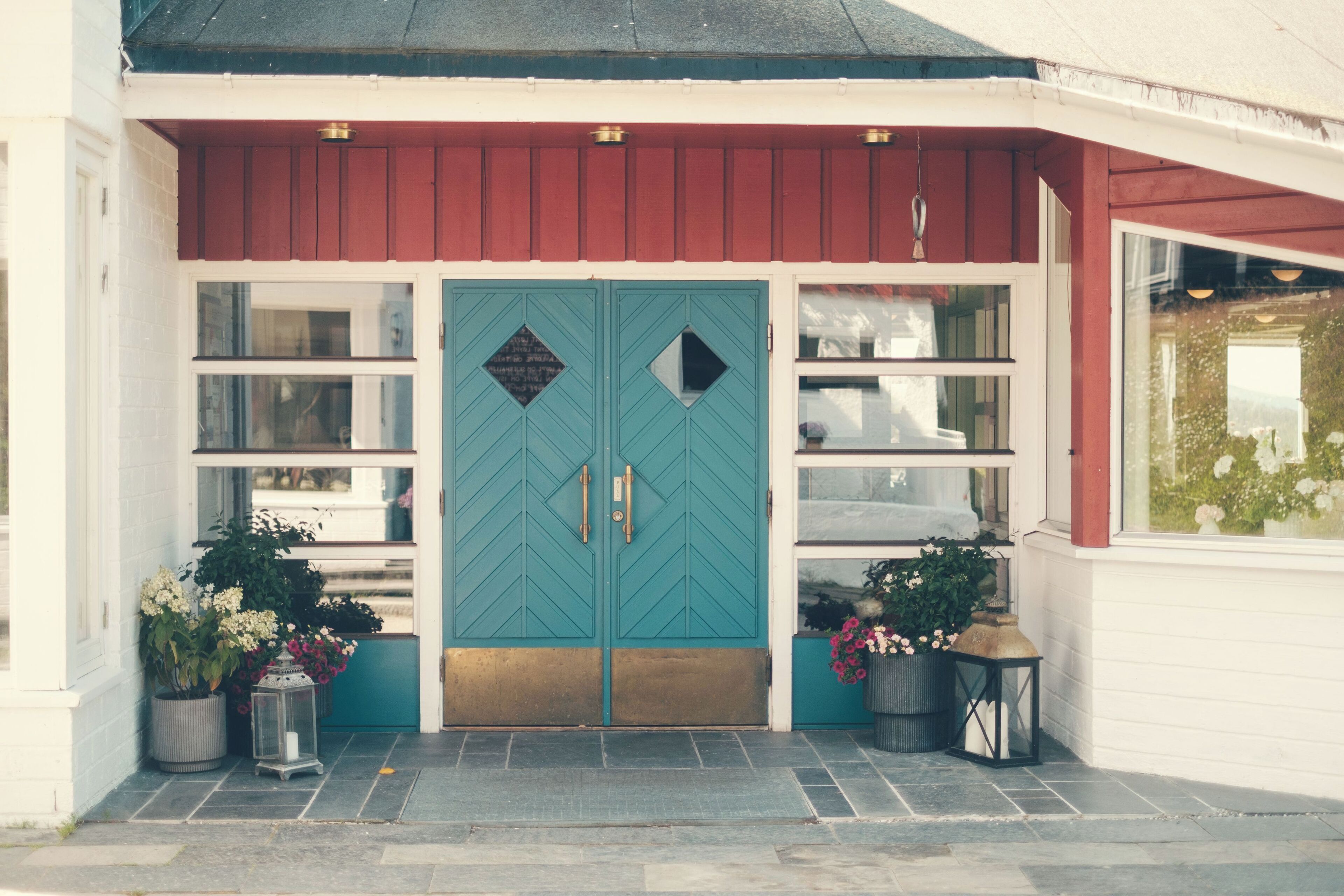 A building with a teal double door, white walls, and a red roof. There are potted plants and a lantern on the ground.