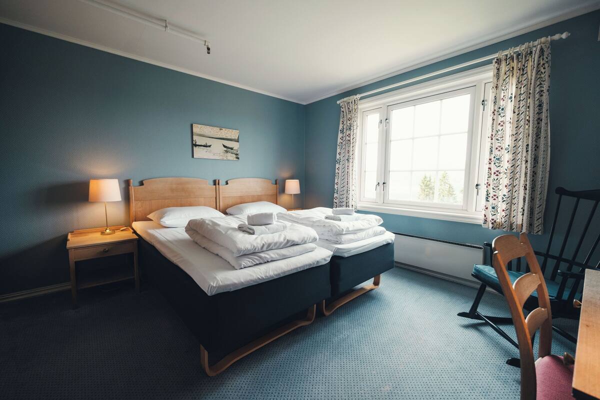 A hotel room with a double bed, a nightstand with a lamp, a wooden chair, and a window with curtains.