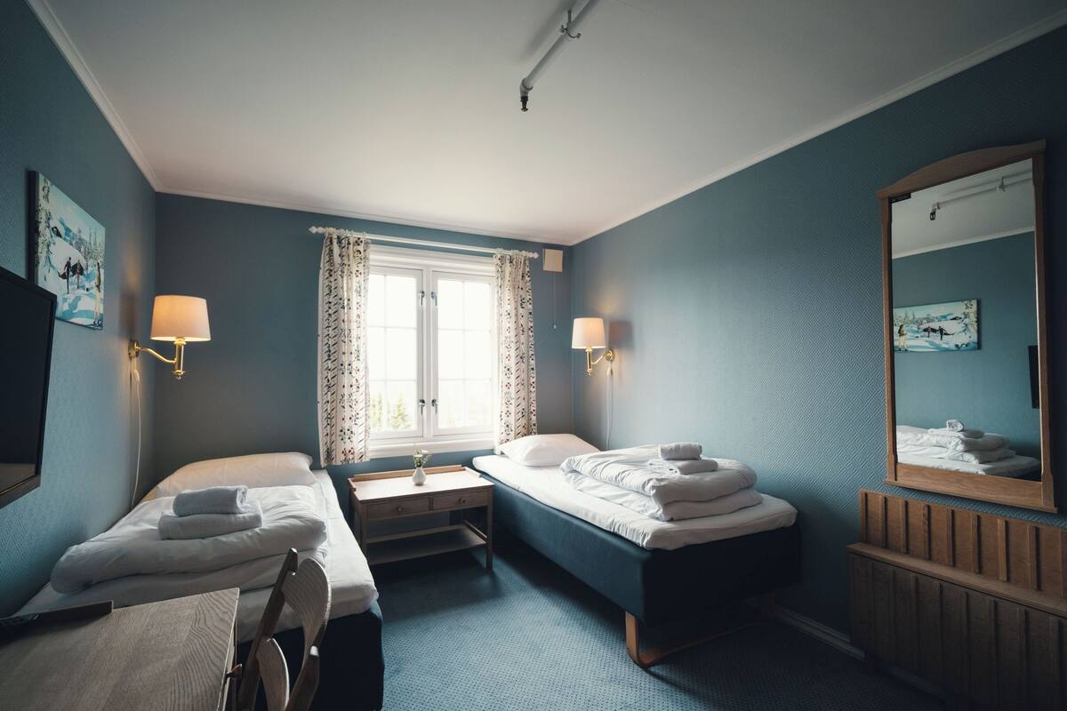 A hotel room with two single beds, a small table, a chair, a window with curtains, and a mirror on the wall.