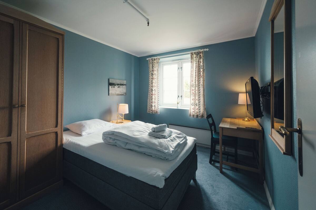A bedroom with a bed, a desk, a chair, a wardrobe, and a window with curtains.