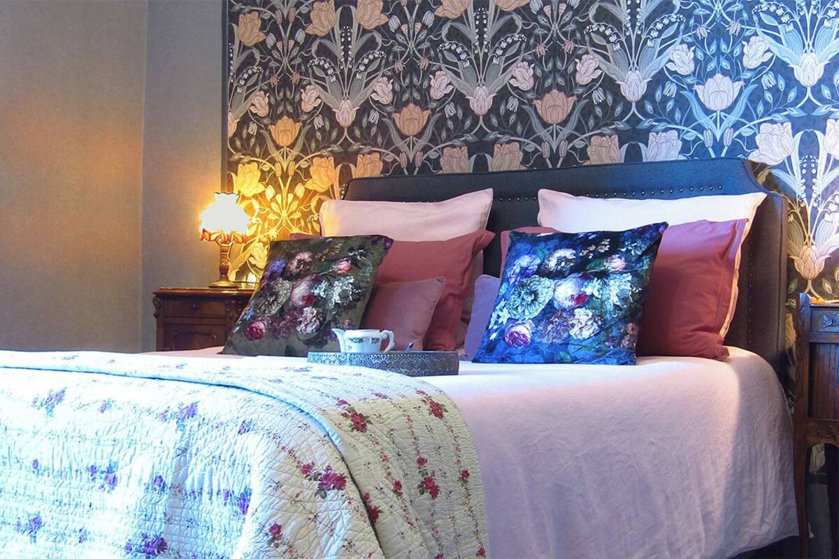 A neatly made bed with floral-patterned pillows and a quilt, a bedside table with a lamp and a cup, and a wall with a floral wallpaper pattern.