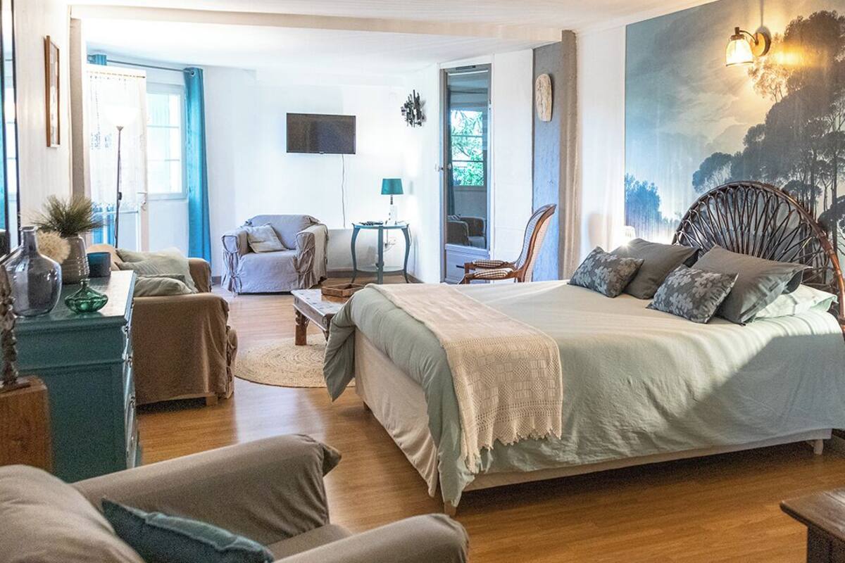 A spacious bedroom with a large bed, a sitting area with a chair and a table, and a view of a forest through the window.