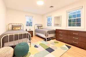 Ein Schlafzimmer mit zwei Metallbetten, einer Holzkommode, einem farbenfrohen Teppich und großen Fenstern mit Jalousien.