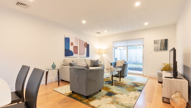 A modern living room with a grey sofa, a coffee table, and a TV. There is a large window with a view of a fence.