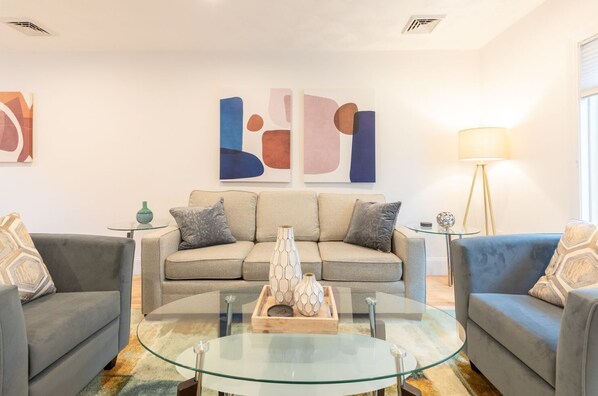 A modern living room with a beige sofa, two grey armchairs, a glass coffee table, and abstract wall art. - Private Home Near Kendall Sq (Cambridge)